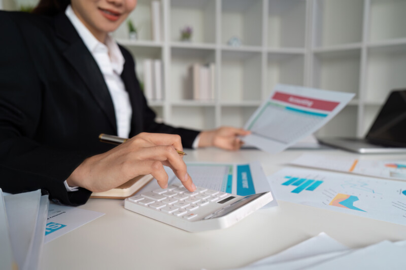 Professional businesswoman in a suit analyzing financial documents and using a calculator at a desk in a modern office.